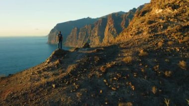 Sırt çantalı adam, akşam vakti gökyüzünün arka planında, okyanus kıyısında, dağın tepesinde duruyor. Sırt çantalı turistlerin arka hava görüntüsü kayalık kayalıkları küçümsüyor. Tenerife Kanarya Adası İspanya.