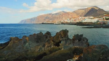 Kayalık kıyı şeridinin arka planında şehir manzarası olan derin okyanus sularının insansız hava aracı görüntüleri. Tenerife, Atlantik Kanarya Adaları. Güneşli yaz günü.