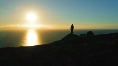 İnsan silueti uçurumun kenarında durur ve gün batımında okyanusa bakar. Güneş denizdeki su yüzeyine dokunur. Hava aracı uçuşu. Los Gigantes Tenerife Kanarya Adaları İspanya Avrupa. Süre