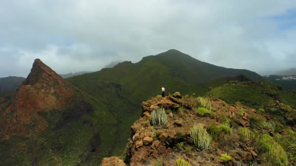 山の中でショット女性バックパッカーのハイキングの周りの空中ドローン 少女は崖の端をクロール登る 観光客は自然火山の風景を楽しむテネリフェカナリア諸島スペインヨーロッパ ロック リスコ — ストック動画