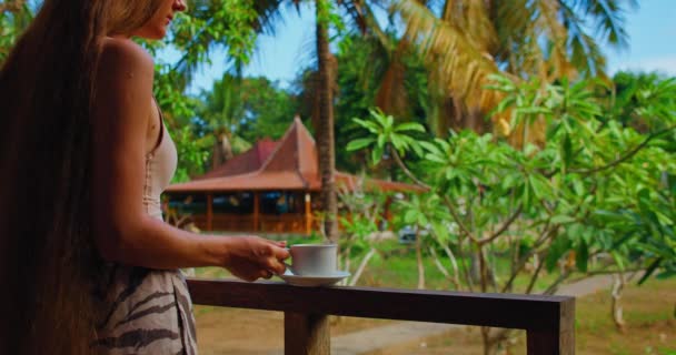 美しいスレンダーな女性は 伝統的なバリの建物と緑の熱帯の自然とバルコニーで朝のコーヒーや紅茶を飲んでいます 旅行の女の子 ヌサペニダ島インドネシア 近くの手と体 — ストック動画
