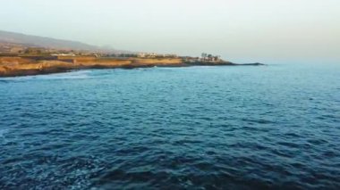 Mavi sakin okyanus dalgaları, plajda volkanik kayalar bulunan turistik şehrin yakınındaki kayalık sahil şeridine çarpar. Volkanik Tenerife kıyısındaki tatil beldesi. İspanya. Kanarya Adaları.