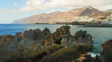 Kayalık kıyı şeridinin arka planında şehir manzarası olan derin okyanus sularının insansız hava aracı görüntüleri. Tenerife, Atlantik Kanarya Adaları. Güneşli yaz günü.