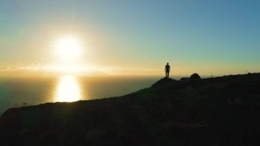 İnsan silueti uçurumun kenarında durur ve gün batımında okyanusa bakar. Güneş denizdeki su yüzeyine dokunur. Hava aracı uçuşu. Los Gigantes Tenerife Kanarya Adaları İspanya Avrupa. Süre