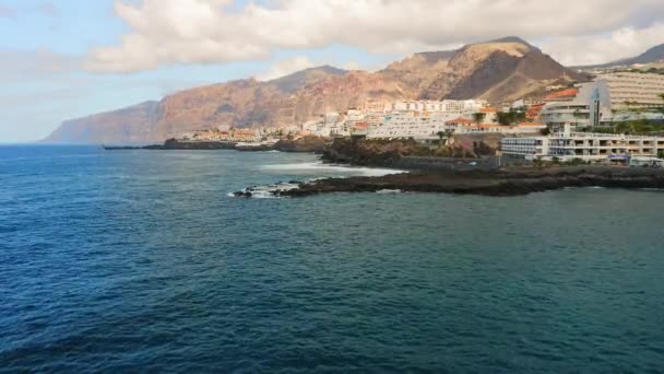 在巨大岩石的背景下 平静的海浪冲击着旅游城市附近的海岸线 夏日晴天 圣地亚哥港的城市景观 Los Gigantes悬崖Tenerife加那利群岛 — 图库视频影像