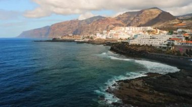 Tenerife volkanik adasının kıyısındaki tatil köyü. Sakin okyanus dalgaları, turizm kenti yakınlarındaki sahil şeridine büyük kayalıklara çarpıyor. Yaz güneşli bir gün. Puerto de Santiago şehir manzarası. Los Gigantes