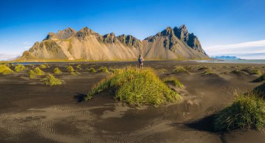 İzlanda 'nın güneydoğu kıyısındaki Stokksnes burnundaki siyah kum tepeciğindeki kadın ve İzlanda' nın Vestrahorn yaz manzarası. Havadan panoramik çekim.