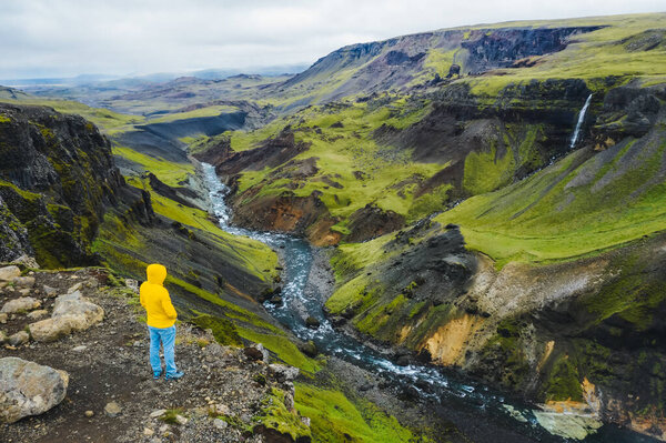 Man in yellow jacket enjoying Highlands of Iceland. River Fossa stream in the Landmannalaugar canyon valley. Hills and cliffs are coverd by green moss.
