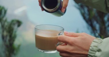 Woman hands pouring coffee from reusable container into a glass cup on nature background. Close-up slow motion. Mountain top overlooking the coast. Anaga rural park. Forest landscape. Tenerife Canary