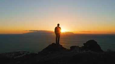 Person standing on top of rock with epic mountain viewpoint colorful sunset and endless ocean horizon. Drone aerial landscape. Flight aroud the man. Tenerife Canary Islands Spain Europe.