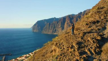 Person standing on top of rock with epic mountain viewpoint colorful sunset and endless ocean horizon. Drone aerial landscape. Mid-adult man. Los Gigantes. Tenerife Canary Islands Spain Europe.