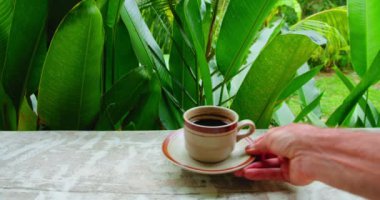 Waiter hand serving a cup of black coffee luwak on countertop. Man bringing coffeinated chocolate drink. Outdoor beverage espresso doppio americano. Wooden table in green lush tropical forest or