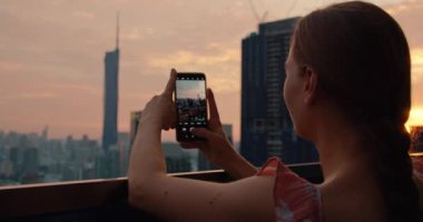 Çekici esmer kadın elinde bir telefon tutuyor ve akşam şehrinin fotoğrafını çekiyor. Terasta modern yüksek binalar var. Seyahat konsepti.