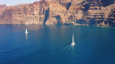 Büyük yüksek dağlar ve derin mavi gök mavisi okyanus sularında sürüklenen yalnız beyaz bir yat. Parlak bir öğleden sonra küçük bir teknenin havadan görüntüsü. Tenerife Kanarya Adası İspanya. Doğa manzarası.