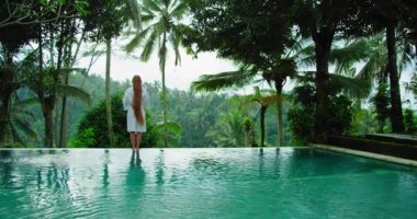 Girl nymph meets the morning stretching by infinity pool enjoying luxury lifestyle on exotic summer vacation with view of tropical jungle forest in foggy morning. Rear view of long-haired woman in