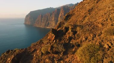 Warm sunset light at Los Gagantes cliffs with deep calm ocean. Flight along high rock crag mountains Tenerife Canary Islands Spain Europe. Aerial footage. Nobody.