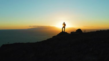 Silhouette of hiker man standing on mountain top at sunset. Boy looking at deep ocean and orange dark sky. Adventure freedom exploration of Planet Earth. Tenerife Canary Islands Spain Europe.