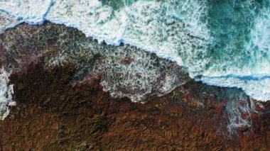 Soothing view of ocean waters washing volcanic rocky rough beach, waves crashing on coastal rocks and making white foam. Transparent shallow emerald azure sea shore. Volcanic lava shoreline. Aerial