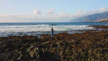 Beautiful aerial shot of female stands on rocky ocean and watching wide powerful foaming waves. Rear view of lonely woman contemplates natural landscape. Tenerife Canary Islands Spain Europe.