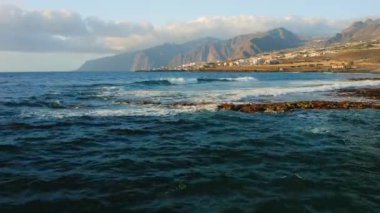 Blue calm ocean waves crash on rocky coastline near tourist city with volcanic rocks in the beach. Resort cityscape on the coast of Island Tenerife. Canary Spain. Aerial static footage. Nobody.