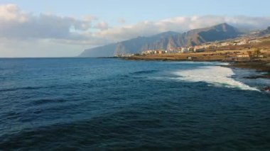 Blue calm ocean waves crash on rocky coastline near tourist city with volcanic rocks in the beach. Resort cityscape on the coast of Island Tenerife. Canary Spain. Aerial drone footage. Nobody.
