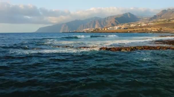 青い穏やかな海の波は ビーチに火山岩がある観光都市の近くの岩だらけの海岸線で衝突します テネリフェ島の海岸沿いのリゾートの街並み スペインのカナリア 空中静止画 — ストック動画