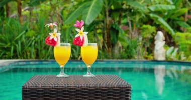 Havuz kenarında çiçekli iki sarı kokteyl suyu. Yüzme havuzu mavi su arka planı. Yeşil yemyeşil yağmur ormanları, palmiye ağaçları. Tatilde. Yavaş çekim videosu.