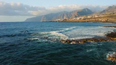 Blue calm ocean waves crash on rocky coastline near tourist city with volcanic rocks in the beach. Resort cityscape on the coast of Island Tenerife. Canary Spain. Aerial drone footage. Nobody.