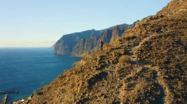 Person standing on top of rock with epic mountain viewpoint colorful sunset and endless ocean horizon. Drone aerial landscape. Mid-adult man. Los Gigantes. Tenerife Canary Islands Spain Europe.