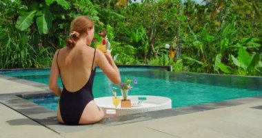 Woman relaxing drinking welcome drink in pool. Luxury vacation in modern resort tourist spot. Traveler sitting and sunbathing near swimming basin. Refreshment and enjoy tropical view with green nature