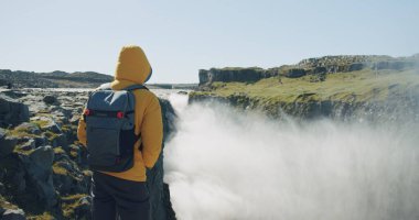 Sarı ceketli ve sırt çantalı bir adamın uçurumun kenarında dikilip İzlanda 'daki Detifoss şelalesine bakması ve eğlenmesi..