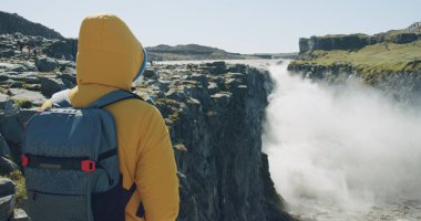 Sarı ceketli ve sırt çantalı bir adam uçurumun kenarında dikilip İzlanda 'daki Detifoss şelalesine bakıyor ve eğleniyor..