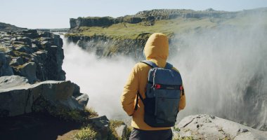 Sarı ceketli ve sırt çantalı bir adam uçurumun kenarında yürüyüp İzlanda 'da Detifoss şelalesinin keyfini çıkarıyor..