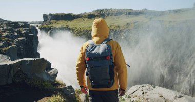 Sarı ceketli ve sırt çantalı bir adam uçurumun kenarında yürüyüp İzlanda 'daki Detifoss şelalesine bakıyor..
