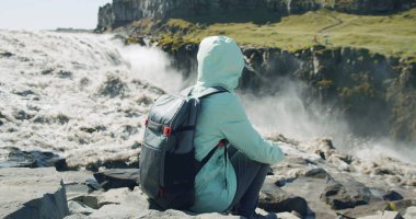 Uçurumun kenarında oturan kadın Detifoss Şelalesi 'ne dökülen gölün arka planındaki İzlanda' ya odaklanmış..