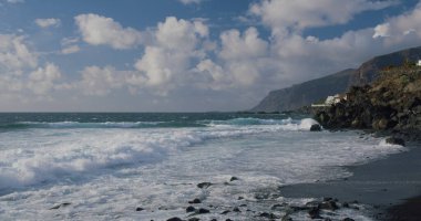 Tenerife, Kanarya Adası, Puerto de Santiago, Playa veya plaj la Arena. Kara kumlu sahil manzarası, Atlantik Okyanusu. Ön planda inanılmaz köpüklü dalgalar var. Arka planda Los Gigantes 'in dev uçurumları var. Gökyüzünde güzel bulutlar. Kimse..