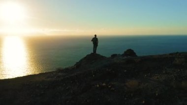 Silhouette of hiker man standing on mountain top at sunset. Boy looking at deep ocean and orange dark sky. Adventure freedom exploration of Planet Earth. Tenerife Canary Islands Spain Europe.