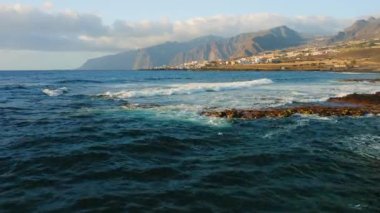 Blue calm ocean waves crash on rocky coastline near tourist city with volcanic rocks in the beach. Resort cityscape on the coast of Island Tenerife. Canary Spain. Aerial static footage. Nobody.
