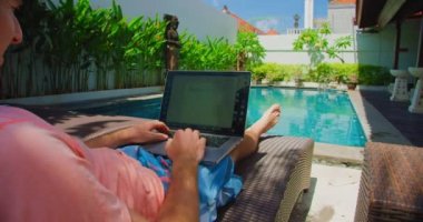 Adult man relaxing on a deck chair by the pool and study or working on PC, computer software. Male hands type on a laptop keyboard while resting outside, summer vacation background. Remote working.