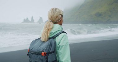 Siyah kumsalda yürüyen, yeşil yağmurluklu bir kadın. Reynisfjara 'nın deniz kenarındaki uçurum oluşumlarının keyfini çıkarıyor..