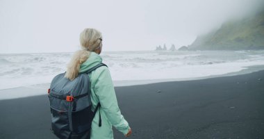 Siyah kumsalda yürüyen, yeşil yağmurluklu bir kadın. Reynisfjara 'nın deniz kenarındaki uçurum oluşumlarının keyfini çıkarıyor..