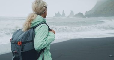 Siyah kumsalda yürüyen, yeşil yağmurluklu bir kadın. Reynisfjara 'nın deniz kenarındaki uçurum oluşumlarının keyfini çıkarıyor..