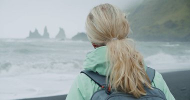 Siyah kumsalda yürüyen, yeşil yağmurluklu bir kadın. Reynisfjara 'nın deniz kenarındaki uçurum oluşumlarının keyfini çıkarıyor..