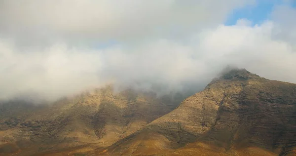 stock image Peaks of massive lifeless mountains covered by thick white fog clouds at sunrise. Outdoor adventure road trip travel to Fuerteventura, Cofete sand beach. Nobody. High quality footage 4k.