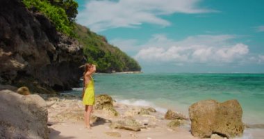 Happy cheerful girl in dress enjoys vacation on white sand beach at summer sunny day. Barefoot attractive woman spend time on ocean, waves foaming. Smiling lady have fun on seashore, beautiful nature.