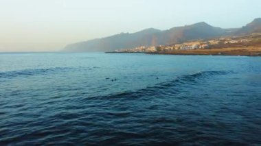 Blue calm ocean waves crash on rocky coastline near tourist city with volcanic rocks in the beach. Resort cityscape on the coast of Tenerife. Spain. Canary Islands.