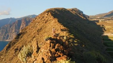 Hiker girl relaxing on rocky mountain top enjoying sunset nature in travel, wilderness trail. Lonely woman traveler in hiking contemplates ocean view. Healthy lifestyle concept. Tenerife Canary