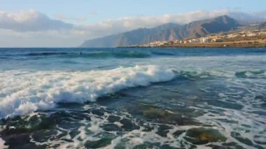 Atlantik Okyanusu 'nun boş kayalık volkanik plajı yumuşak sabah ışığında kırılan dalgaların üzerinde alçaktan uçuyor. Deniz köpüğü. Arka planda tatil köyü, şehir manzarası. Tenerife Kanarya Adaları İspanya Avrupa.