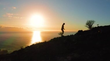 Bir kızın silueti, okyanus suyunun üzerinde gün batımını izleyerek yokuş yukarı tırmanıyor. Dağın kenarında yürüyen bir kadının havadan görünüşü. Arka planda büyük güneş var. Alacakaranlık, gece doğası. Tenerife Kanaryası