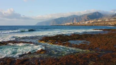 Okyanus dalgaları sahilin kayalık kıyılarına çarpıyor. Tuzlu deniz suyu köpüğü. Arka planda tatil köyü. Rocky volkanik Tenerife Adası. Şehir manzarası. İspanya Avrupa.